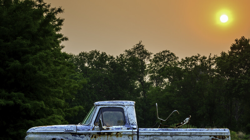 Abandoned pickup truck in overgrown field at golden hour with dense forest backdrop.
