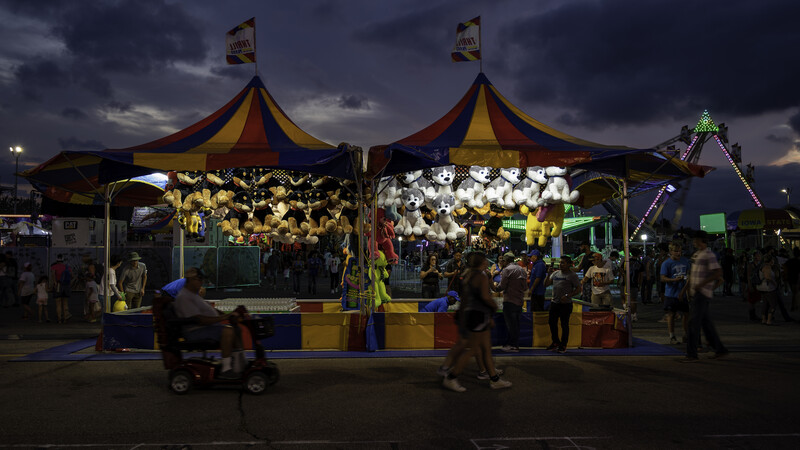 Two illuminated carnival game booths with yellow and red striped tents at dusk, surrounded by crowds and fairground attractions.
