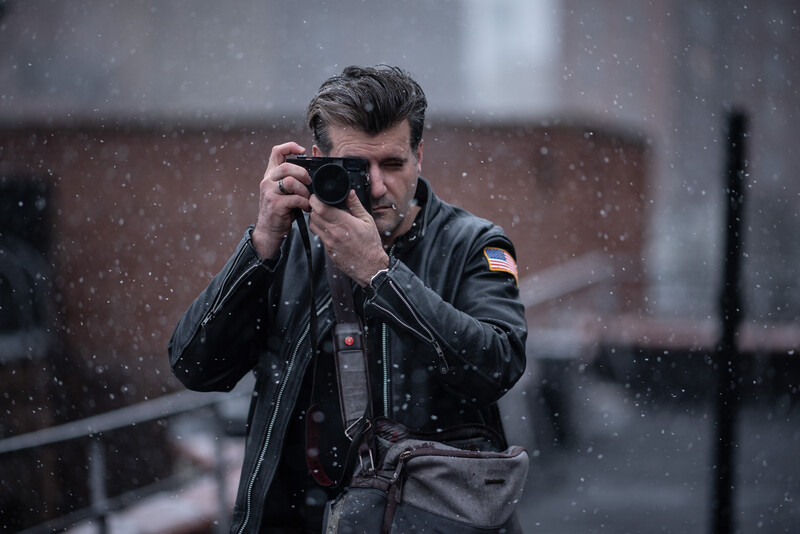 Man in black leather jacket holding camera up to photograph in falling snow.