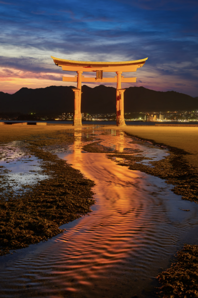 Illuminated torii gate reflected in wet sand at dusk with mountains and lights in background.