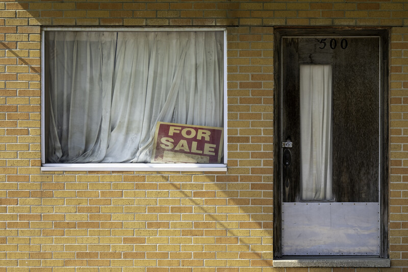 Abandoned residential storefront with "For Sale" sign in window and curtains visible behind glass.