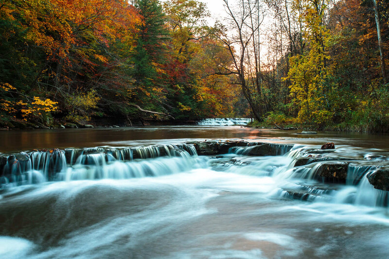 Cascading waterfall flowing through a forested river surrounded by autumn foliage.