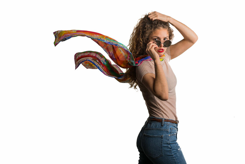 Woman in sunglasses with colorful patterned fabric flowing behind her against white background.