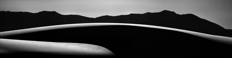 Curved white sand dune with sharp ridge line against dark silhouetted mountains under overcast sky.