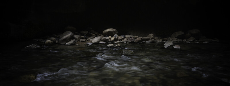 Dark stream flowing over smooth rocks beneath a large boulder overhang at night.
