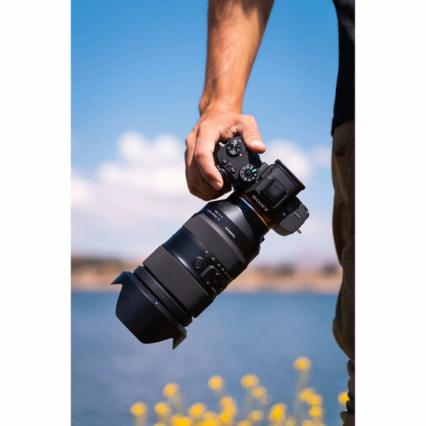 Photographer holding a telephoto lens and camera body against a blue sky and water backdrop.