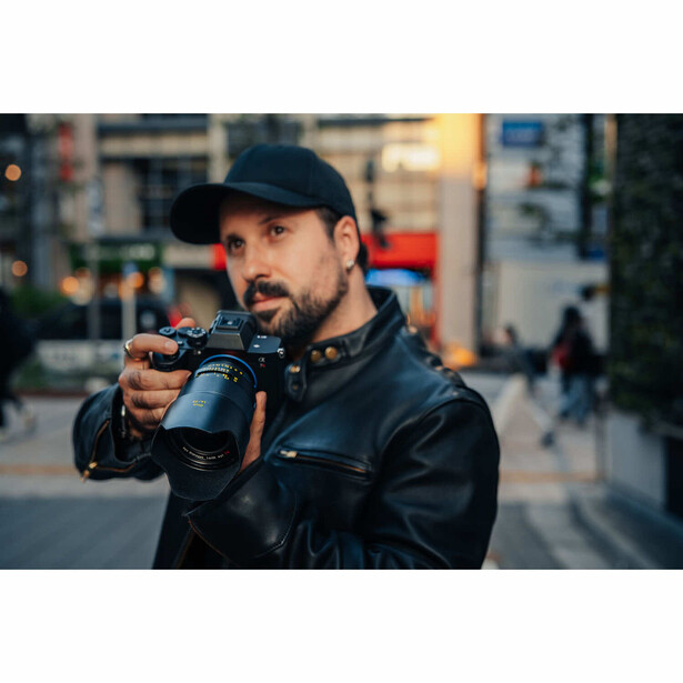 Male photographer holding a mirrorless camera with telephoto lens in urban street setting.