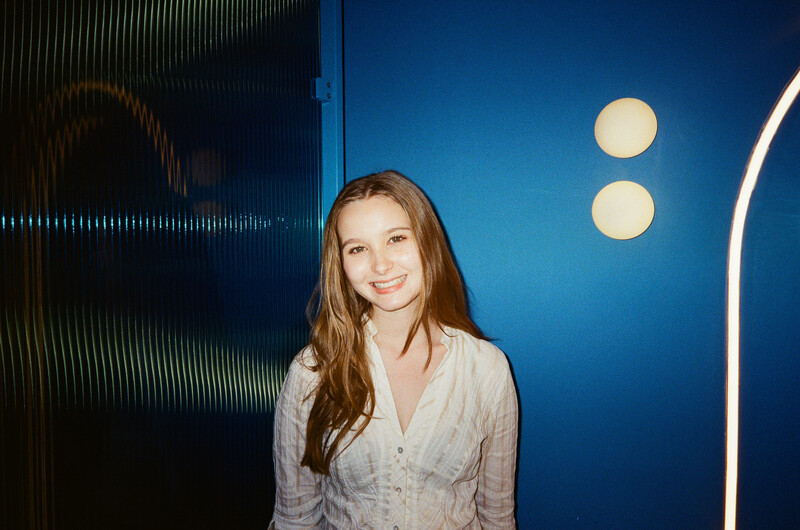 Woman with long brown hair smiling at camera against blue backdrop with modern lighting.