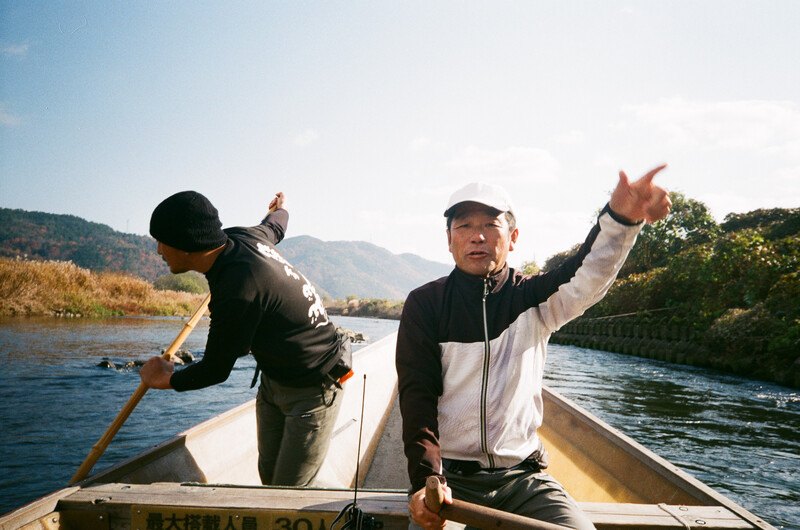 Two men in a small boat on a river with mountains visible in the background.