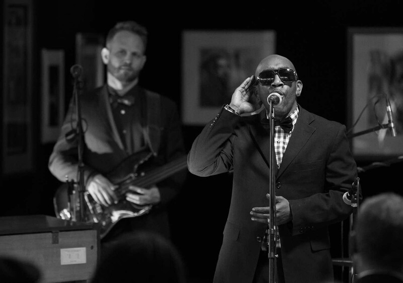 Musician in sunglasses and suit singing at microphone with guitarist in background on stage.
