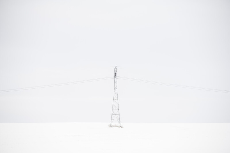 A solitary electrical transmission tower stands in heavy fog or snow, with power lines extending into the mist.