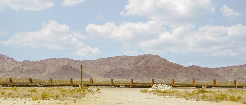 Abandoned railroad flatbed car on rusted tracks in an arid desert landscape with mountains in the background.