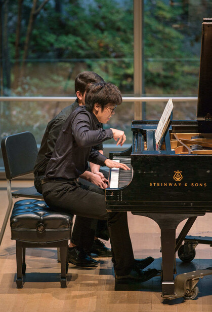 Young pianist performing at a grand piano during a recital with large windows and forest backdrop.