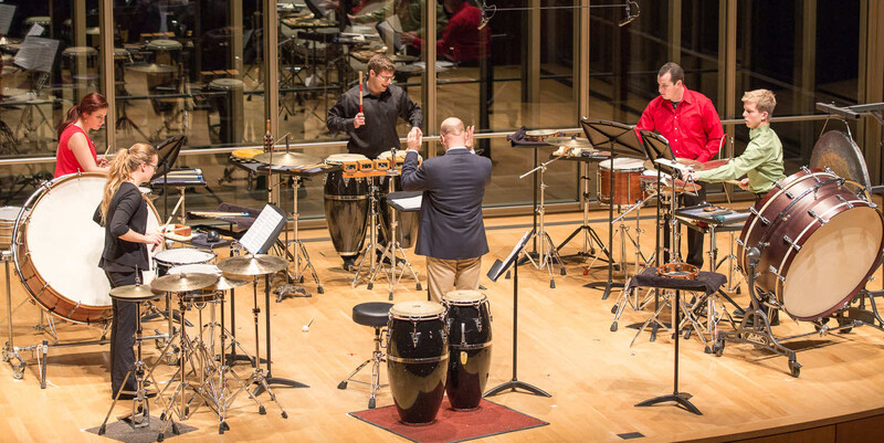 Six musicians performing on a wooden stage with various percussion instruments arranged in a circle.