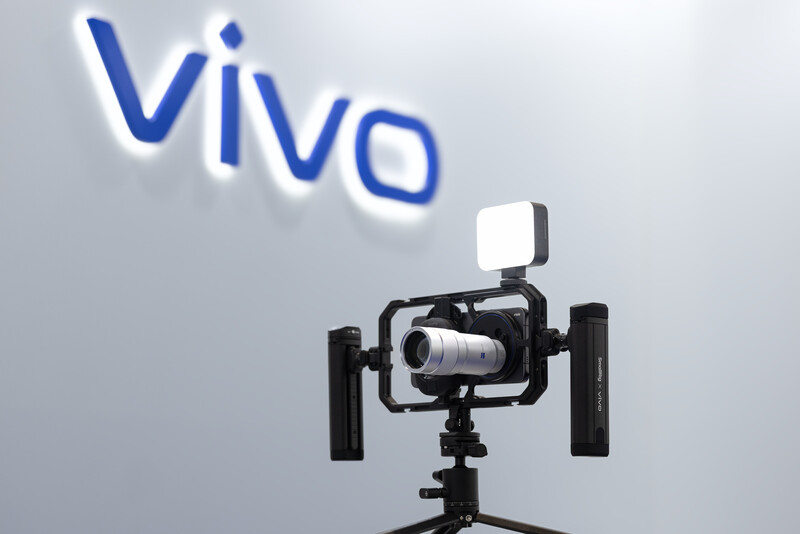 Professional video camera rig with dual side handles, fill light, and lens mounted on tripod under Vivo brand signage.