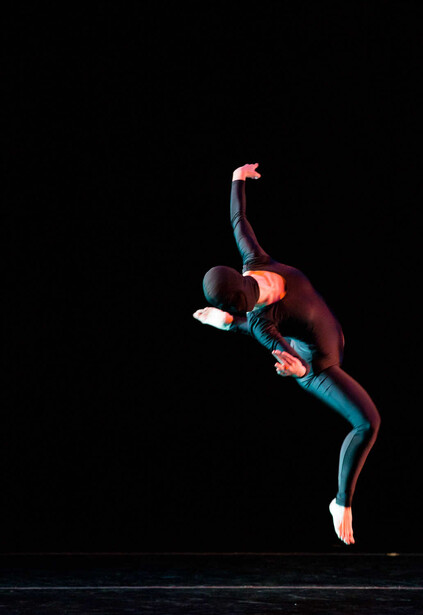 Dancer mid-leap against black background, body arched with one leg extended and arms raised.