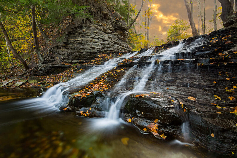 Cascading waterfall flowing over layered slate rocks with autumn leaves scattered across the surface.
