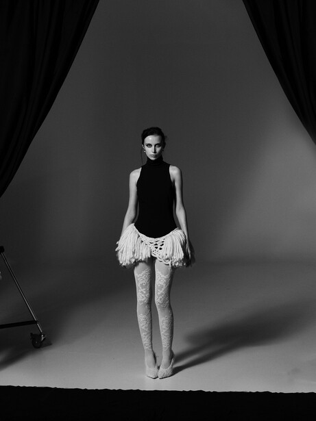 Woman in sleeveless black top and white lace skirt standing on gray seamless background under studio lighting.