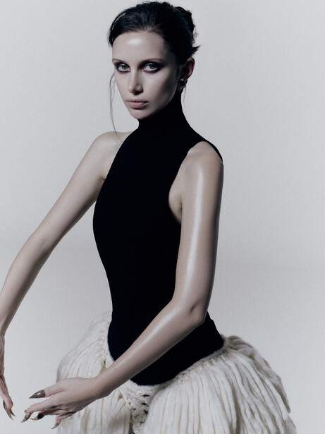 Dancer in black sleeveless leotard and white tulle skirt, posed in profile against neutral background.