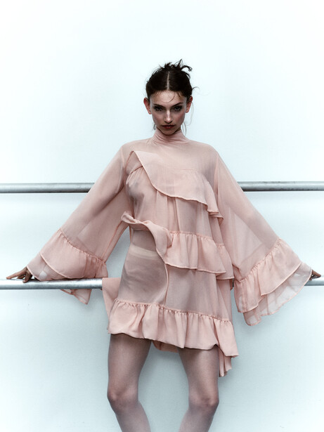 Model wearing layered pink ruffle dress against white wall with horizontal metal rail.