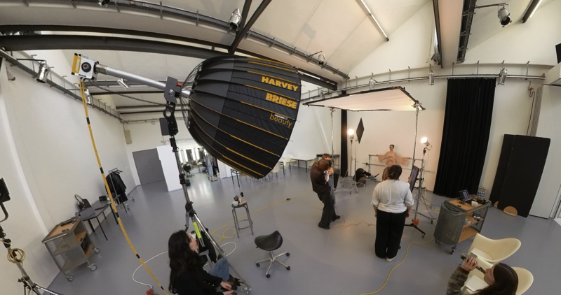 Professional photography studio with large soft light modifier suspended overhead and crew setting up for a shoot.