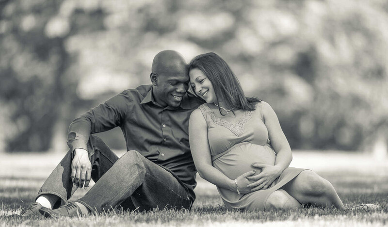 Expectant couple sitting on grass, man with arm around pregnant woman smiling at camera.