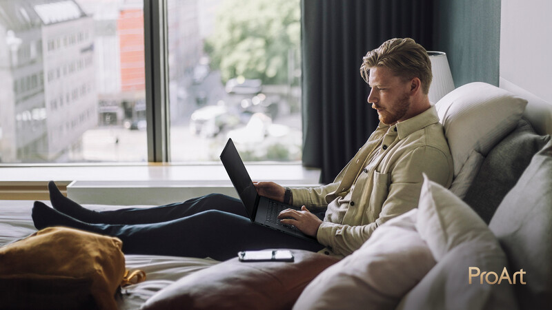 Man working on laptop while reclining on a bed near a large window overlooking a city street.