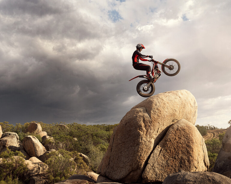 Dirt bike rider performing a wheelie on a large boulder against a dramatic cloudy sky.
