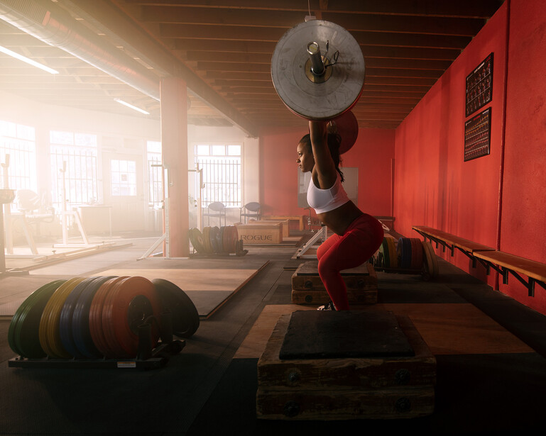 Athlete performing barbell overhead press in a gym with red walls and natural light streaming through windows.