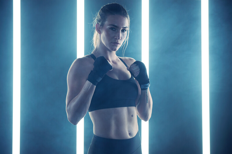 Athletic woman in boxing stance wearing hand wraps against blue neon vertical light panels.