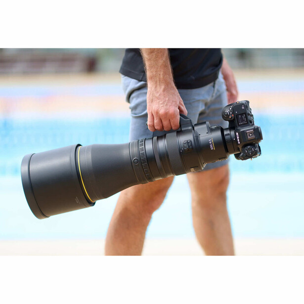 Photographer holding a large telephoto lens with camera attached, outdoors by a swimming pool.