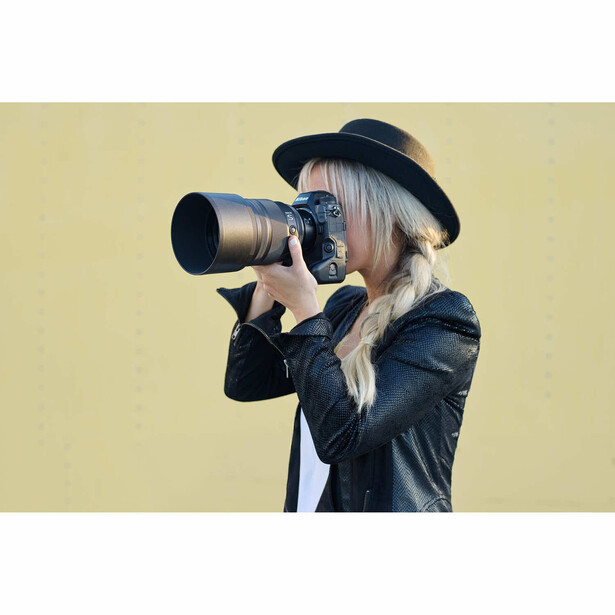 Photographer with long blonde hair and black hat holding telephoto lens camera to eye against yellow background.