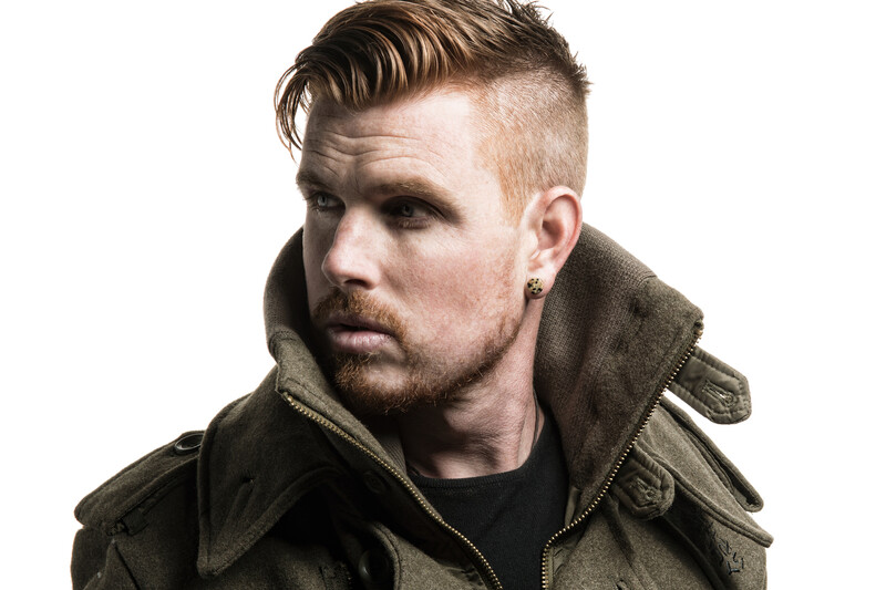 Male portrait with styled undercut hairstyle wearing olive-green jacket against white background.