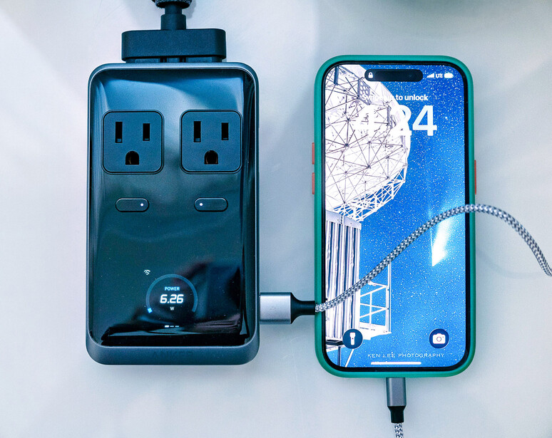 Smartphone charging from a dual-outlet power strip displaying time on its screen.