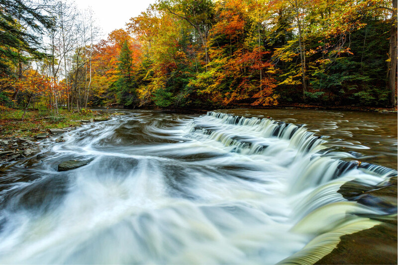 Cascading waterfall with silky motion blur flowing over stepped rocks through autumn forest.