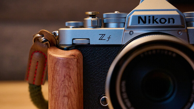 Nikon Z f mirrorless camera with wooden hand grip and red leather strap against neutral background.