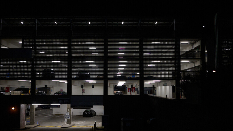 Modern parking garage at night with illuminated storefront windows displaying merchandise and vehicles.