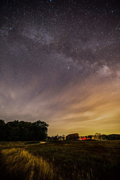 Milky Way galaxy stretches across a star-filled night sky above a darkened rural landscape with silhouetted trees and glowing horizon.