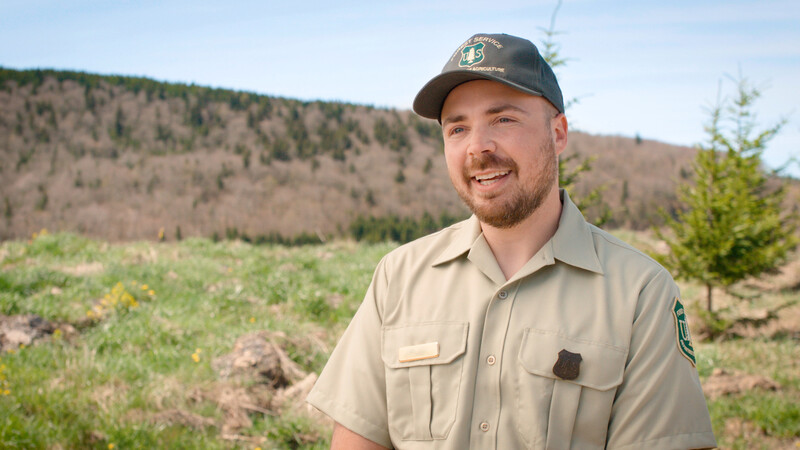 Forest ranger in tan uniform and cap smiling at camera with mountainous terrain and evergreen trees in background.