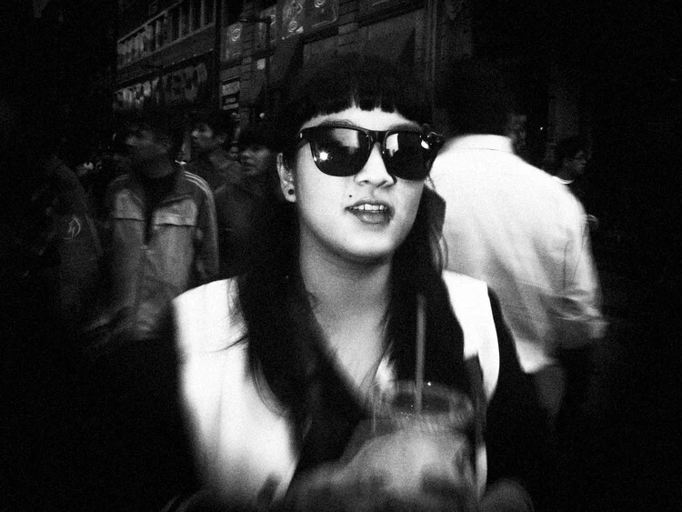 Woman wearing dark sunglasses and white collar in crowded urban setting, shot in black and white.