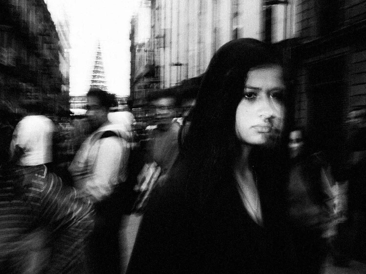 Woman with long dark hair in urban street amid blurred pedestrians and buildings.
