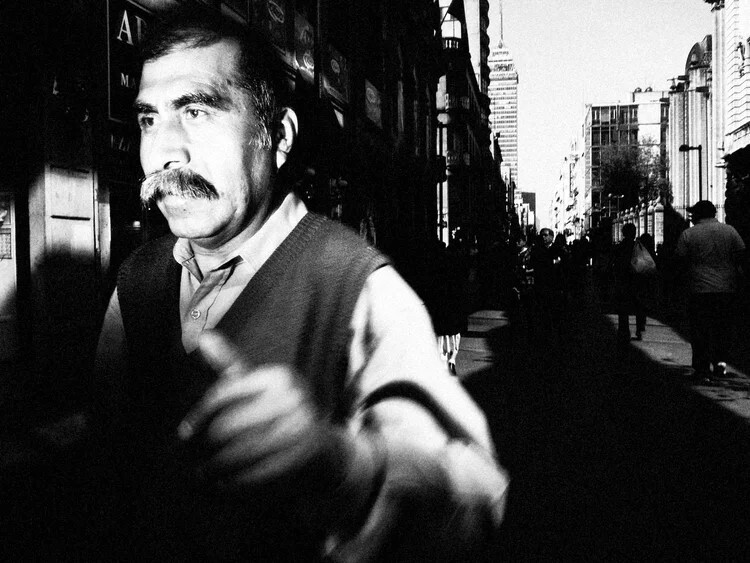 Man with prominent mustache on an urban street, shot in black and white with shallow depth of field.