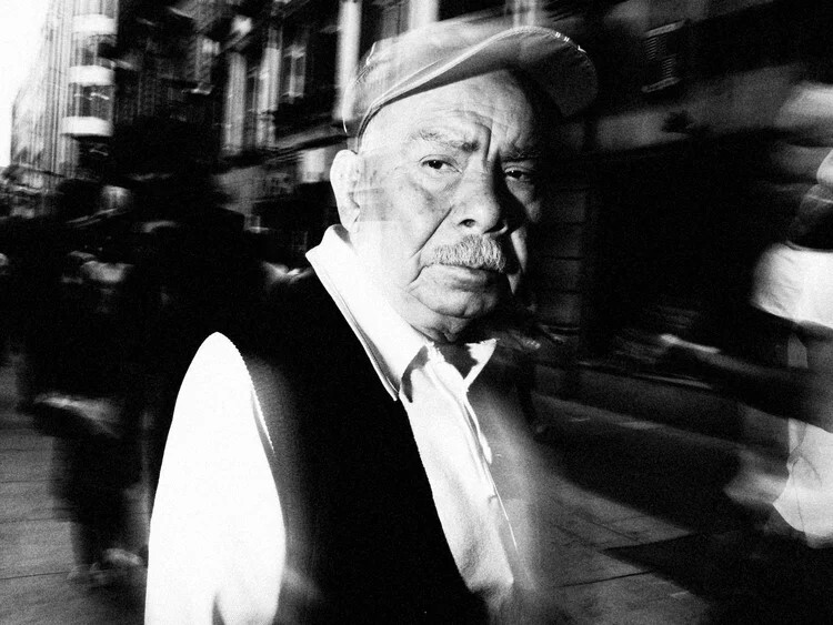 Older man in cap and vest on busy city street with motion blur surrounding him.