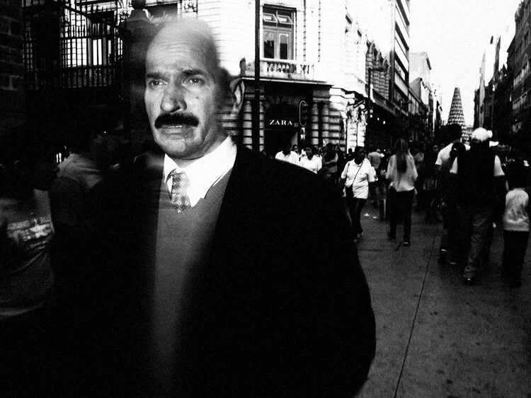Man with distinctive mustache in dark suit on crowded city street, black and white photograph.