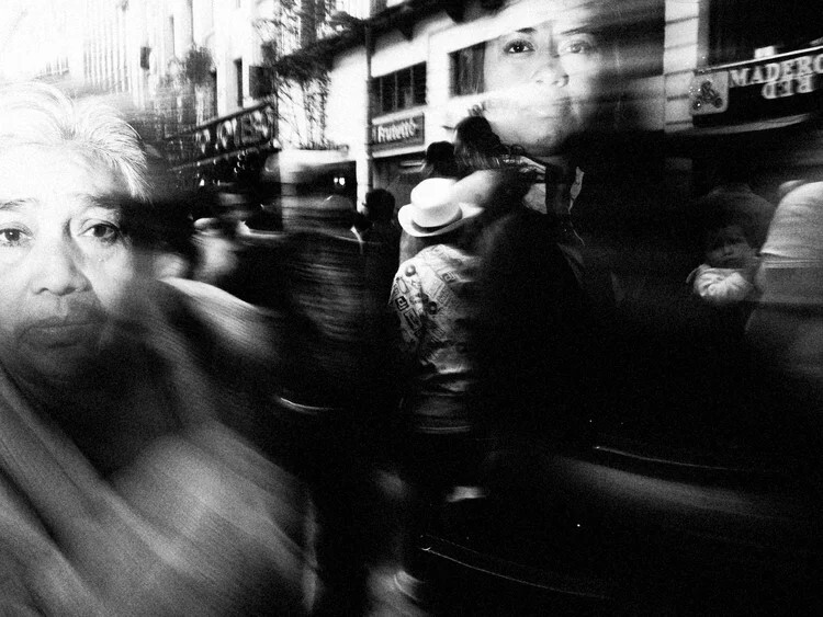 Blurred black and white street scene with multiple figures and storefronts, captured with motion blur.
