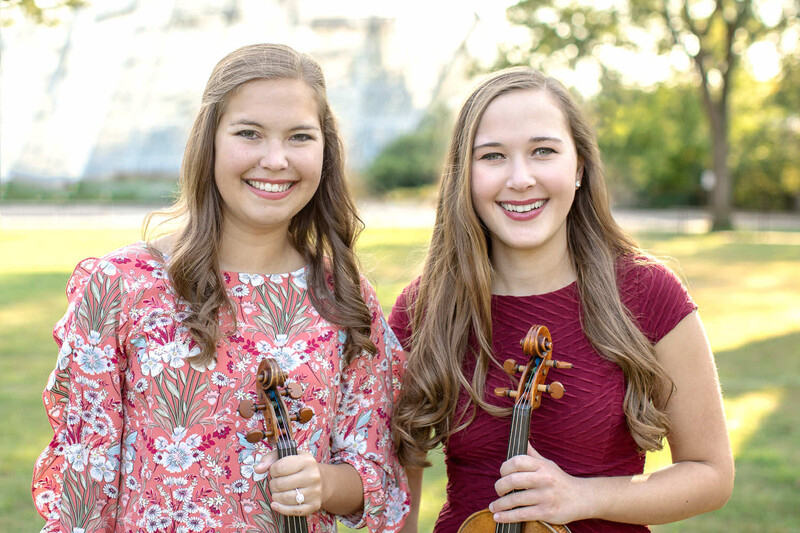 Two young musicians holding violins outdoors in natural daylight.