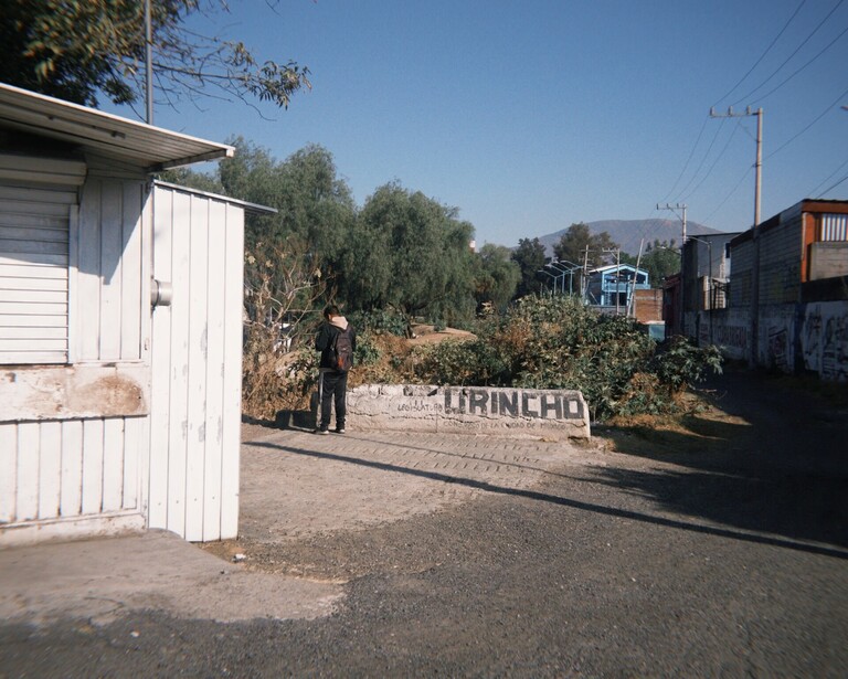 White metal utility structure beside unpaved road in rural settlement with scattered buildings and dry vegetation.