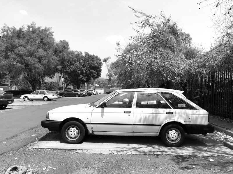 White sedan parked on residential street lined with mature trees and other vehicles.