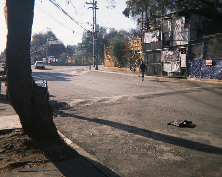 Street scene with long tree shadow cast across asphalt road, residential buildings visible on right side.