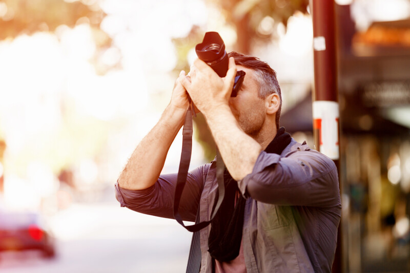 Photographer holding camera up to face against bright, backlit outdoor setting with warm lens flare.